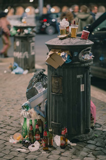 A sanitation worker wearing a high-visibility orange vest and black trousers is seen standing behind the rear of a large red rubbish collection truck, which is parked on the side of a street. The work