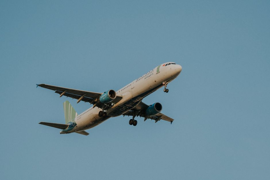 A commercial airplane in mid-flight against a clear blue sky, with its landing gear extended and engines visible beneath the wings. The aircraft features a white fuselage with blue and green accents, 