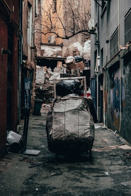 A narrow alleyway cluttered with accumulated rubbish, featuring a large, wheeled black trash bag or container positioned in the foreground, with its fabric appearing crumpled and dirty. Behind it, the