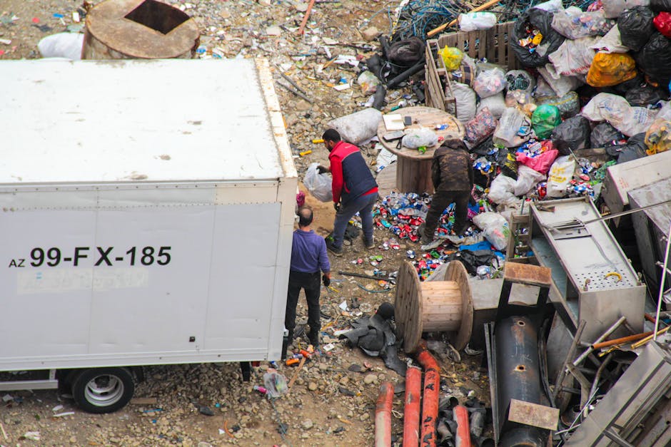An aerial view of a large, untidy landfill site with a mixture of waste materials, including plastic bottles, paper, cardboard, and discarded packaging scattered across a patch of soil. The area is bo