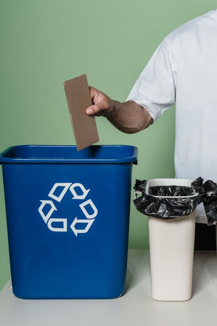 A person's hand, dressed in a white long-sleeved shirt, is seen disposing of crumpled paper into a small, round, light-colored wooden waste bin with a smooth finish. The crumpled paper is beige and wh