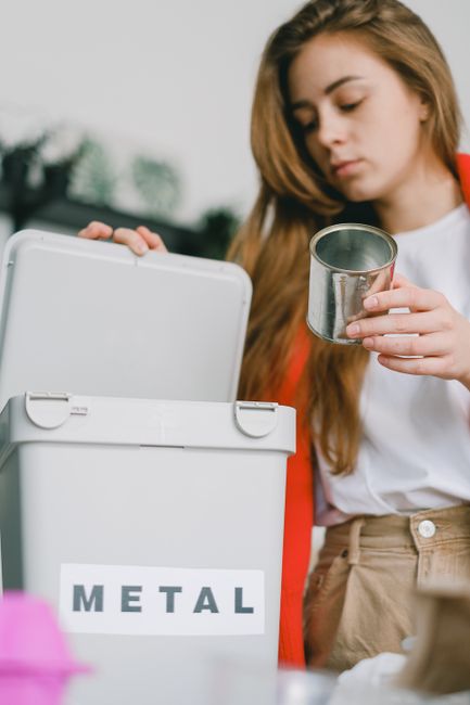 A young woman with long, light brown hair, dressed in a white shirt and beige trousers, stands outdoors in natural daylight. She holds a small, shiny metal container in her right hand, carefully inspe