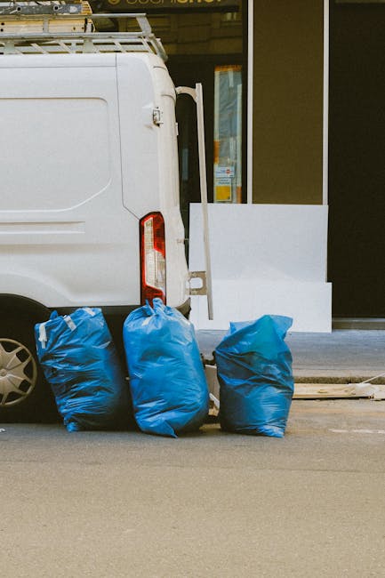 Four large blue plastic rubbish bags, filled and tied at the top, are positioned on the pavement near the rear of a white commercial van, which is parked adjacent to a building with a glass door and w